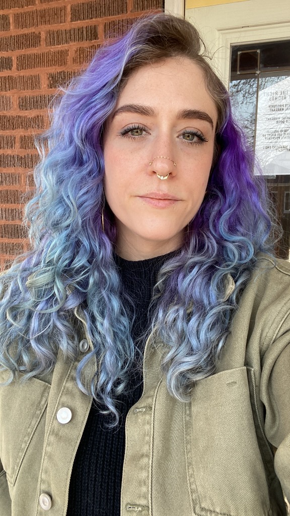 A photo of Alex, a white femme with blue and purple hair slightly smiling. She has an army green denim jacket on and facial piercings
