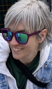 Photo of Al, white non binary person with silver hair looking away from camera and smiling. Wearing purple sunglasses and has on a green sweatshirt, white hoodie, and jean jacket.