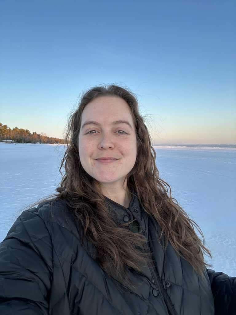 A photo of Emma, a white person with brown wavy hair wearing a black jacket outside during a snowy sunset.