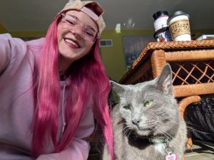 A photo of Elyse (human) and Grey (cat). Elyse has light skin, is wearing glasses, has vibrant pink hair, has nose piercings and is smiling down at the camera. Grey is a grey cat with green eyes looking off to the side.