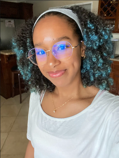 A photo of Angelica, a Brown woman looking into the camera. She has short black hair with bright blue ends, a white headband, large circular black/gold glasses, a gold necklace with a white gem at the end, and a white shirt.